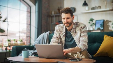 Portrait of Smiling Middle Aged Man Working from Home on a Laptop Computer in Sunny Cozy Apartment. Successful Male Entrepreneur Does Remote for e
