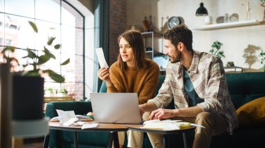 Doing Accounting at Home: Happy Couple Using Laptop Computer, Sitting on Sofa in Apartment. Young Family Filling Tax Forms, Mortgage Documents, Bills
