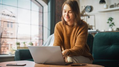 Portrait of Smiling Young Woman Working from Home on Laptop Computer in Sunny Cozy Apartment. Successful Creative Girl Entrepreneur Does Remote for e