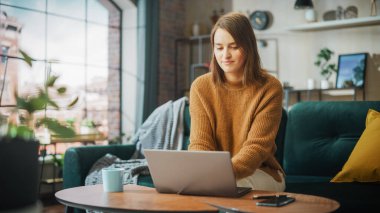 Portrait of Smiling Young Woman Working from Home on Laptop Computer in Sunny Cozy Apartment. Successful Creative Girl Entrepreneur Does Remote for e