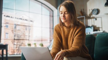 Smiling Young Woman Working from Home on Laptop Computer in Her Sunny Cozy Apartment, Relaxes and Leans Back on a Sofa. Successful Creative Female