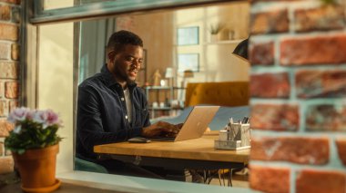 Genç ve yakışıklı erkek dizüstü bilgisayar kullanıyor, apartmandaki oturma odasında oturuyor. Afrikalı Amerikalı İşadamı E-postalara cevap veriyor, İş Planı yazıyor