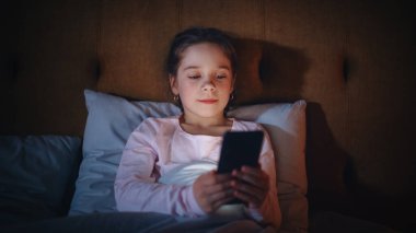 Uykuya dalmadan önce yatakta akıllı telefon kullanan küçük kız. Sosyal medyayı kontrol eden sevimli çocuk, okul programı, Dijital Ebook ödevi okumak, sohbet