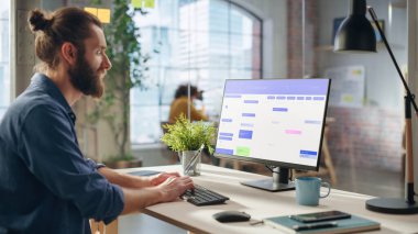 Yakışıklı Uzun Saçlı Yönetici Yaratıcı Ofis Masasında çalışıyor, Masaüstü Bilgisayarını Ekranda Takvim Yazılımı ile kullanıyor. Meslektaşlar