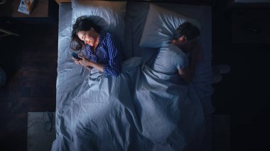 Üst Manzara: Genç Kadın, Erkek Ortağının Uykuya dalmaya çalıştığı Gece Yatağında Akıllı Telefon Kullanıyor. Çift Kavgası, Tartışma. Bağımlılık Dünyası