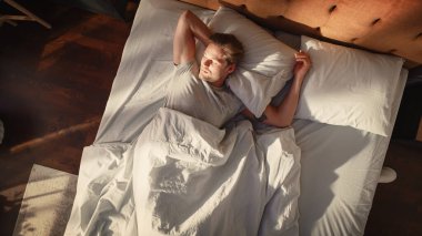 Top View Home: Yakışıklı Genç Adam Yatağında Uyuyor, Üzerine Güneş Parlıyor, Evinde Gözler Açıyor ve Yeni Güneşli Günü Kutluyor. Neşeli Uyanış Bir erkeğin