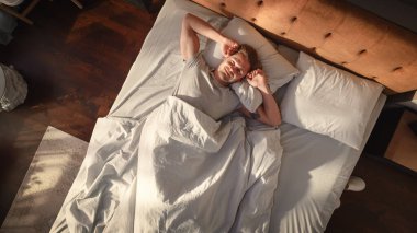 Top View Home: Yakışıklı Genç Adam Yatağında Uyuyor, Üzerine Güneş Parlıyor, Evinde Gözler Açıyor ve Yeni Güneşli Günü Kutluyor. Neşeli Uyanış Bir erkeğin