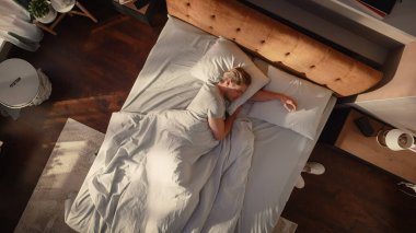 Top View Home: Yakışıklı Genç Adam Şık Bir Yatak Odasında Yatağında Uyuyor, Güneş Üzerine Parlıyor. Günaydın konsept. Huzurlu Uyku ve Rahatlama