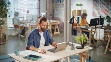 Yaratıcı Ofis Masasında oturan Uzun Saçlı Yakışıklı Sakallı Yönetici. Genç Tarz Adamı Pazarlama Ajansında Dizüstü bilgisayar kullanıyor. Meslektaşlar