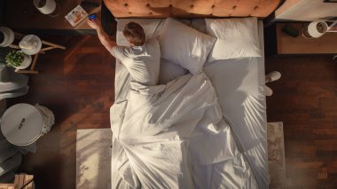 Top View Home: Yakışıklı Genç Adam Yatağında Uyuyor, Smartphone 'da Alarm Saatini Kapatıyor, Evde Yeni Güneşli Günü Kutluyor. Neşeli Uyanış Bir erkeğin