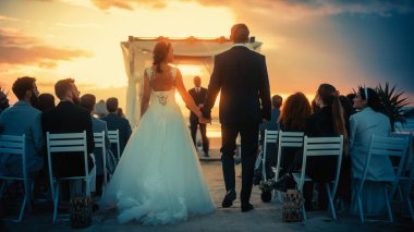 Beyaz Gelinlikli Güzel Gelin ve Siyah Takım Elbiseli Yakışıklı Damat Gün batımında Deniz kenarında bir açık hava seremonisi mekanında