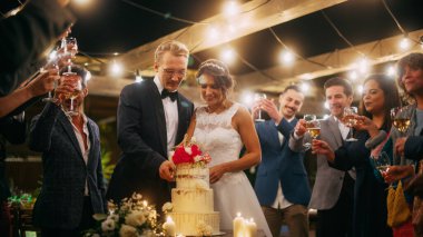 Güzel Gelin ve Damat, Çoklu Etnik Arkadaşlarla Akşam Resepsiyonunda Düğünü Kutluyor. Evli çift yemek masasında dikiliyor, kesiyor.