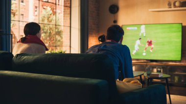 Birkaç futbol hayranı, Stylish Loft Apartment 'da bir kanepede rahatlar, bir spor maçı izlerler. Rahatlamış genç adam ve kadın Favorileri için tezahürat yapıyorlar.