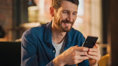 Koltukta oturan yakışıklı bir adamın portresi, Stylish Daire ya da Cafe 'deki akıllı telefonu kullanmak. Yaratıcı Erkek Gülümseyen, Sosyal medyayı kontrol eden ve