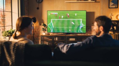 Birkaç futbol hayranı, Stylish Loft Apartment 'da bir kanepede rahatlar, bir spor maçı izlerler. Rahatlamış genç adam ve kadın Favorileri için tezahürat yapıyorlar.