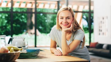 Sarı saçlı, gri V-Neck tişörtlü, Masa 'ya yaslanmış ve gülümseyen güzel yetişkin kadın portresi. Başarılı Kadın