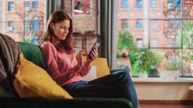 Oturma odasında bir koltukta oturan, rahatlayan ve akıllı telefon kullanan genç ve güzel bir kadın. Yaratıcı Kız Sosyal medyayı kontrol ediyor, Arkadaşlarla sohbet ediyor