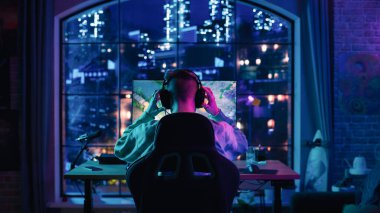 Başarılı bir Stil Oyuncusunun Başının Arkası Kulaklığı Takıyor ve Neon Lit Salonunda Kişisel Bilgisayarda Bir Video Oyunu Oynuyor
