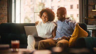 Sevimli siyah çift dizüstü bilgisayar kullanıyor, internetten alışveriş yapıyor, sosyal medyayı kullanıyor. Erkek arkadaş ve kız arkadaş eğleniyor, otururken gülüyor.