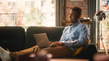 Yakışıklı Afro-Amerikan Adam rahat oturma odasında bir koltukta otururken dizüstü bilgisayarda çalışıyor. Evden çalışan Freelancer. İnternet Taranıyor