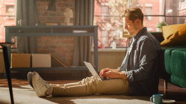 Sunny Loft Oturma Odasında Bir Yerde Oturan ve Dizüstü Bilgisayarı Kullanan Yakışıklı Genç Bir Erkek Portresi. Evden Çalışan Başarılı Adam