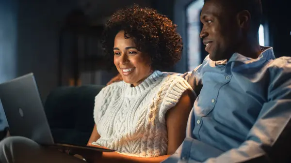 stock image Black Couple Talk with Each Other While Spending Time at Home. They are Sitting on Sofa and Using Laptop for Online Shopping. Man Doing a Tender