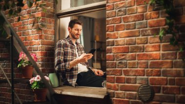Akıllı telefon kullanan ve rahat dairesinin pencere eşiğinde oturan yakışıklı beyaz adam, mutlu ve yakışıklı. Sıradan bir adam tişört giyiyor, internete bakıyor, kontrol ediyor.