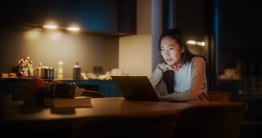 Cozy Kitchen 'da Masada Oturan Güzel Asyalı Kadın ve Geceleri Evde Dizüstü Bilgisayar Kullanmak. Kadın Gülümseyen ve Konuşan