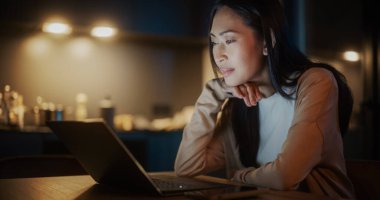 Cozy Kitchen 'da Masada Oturan Güzel Asyalı Kadın ve Geceleri Evde Dizüstü Bilgisayar Kullanmak. Dişi Gülümseyen ve Bakınan