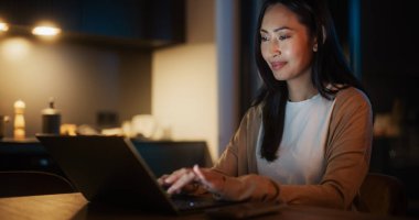 Cozy Kitchen 'da Masada Oturan ve Geceleri Evde Dizüstü Bilgisayarda Yazan Güzel Asyalı Kadın. Dişi Gülümseyen ve Bakınan
