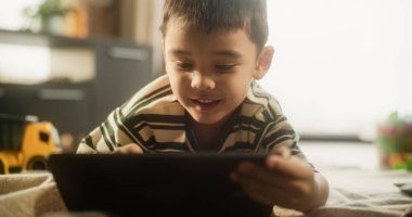Dijital tablet kullanan Asyalı bir çocuğun portresi odasında gülümsüyor. Küçük şirin çocuk hafta sonunun tadını çıkarıyor, eğitici oyunlar oynuyor, resim çiziyor.