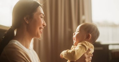 Annelik Sevgisi: Sevecen bir Asyalı Anne 'nin Yakın Çekim Portresi Gülümseyen ve Bebeğiyle Doğal Yatak Odasında Oynayan. Sıcak.