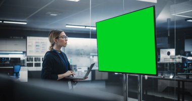 Güzel Yazılım Mühendisi Toplantı Odasında Tek Başına Duruyor, Laptop kullanıyor ve Yeşil Ekran Mock Up ile potansiyeli olan bir TV 'ye bakıyor
