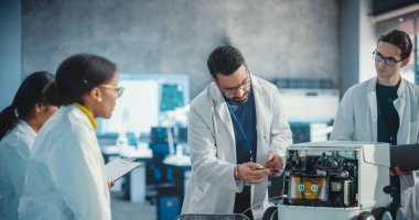 Çeşitli Endüstriyel Robot Uzmanları Mobil Robotlu Masa etrafında toplandı. Laboratuvar Önlüklü Mühendisler Otomatik Yapay Zeka Robotunu Tartışıyor