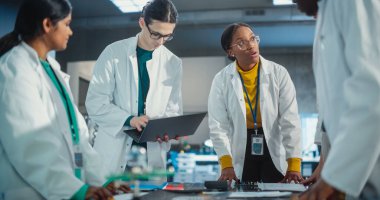 Endüstriyel Robot Uzmanlarından oluşan çeşitli bir ekip bir taslağı tartışmak için bir masanın etrafında toplandı. Laboratuar Önlüklü Mühendisler Laptop ve Tablet Kullanıyor