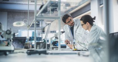 Genç adam laboratuvarda bir fen öğretmeniyle sohbet ediyor. Öğrenci bir işlemci projesi için devre panosunda çalışıyor. Genç Bilgin Konuşuyor