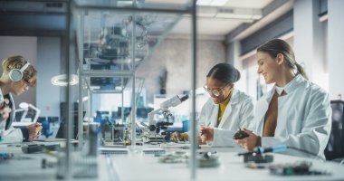 Üniversitedeki Modern Bilim Araştırma Laboratuvarı: Mikroskop kullanan çeşitli beyaz önlüklü genç bilim adamları, Teknoloji için atama tartışması