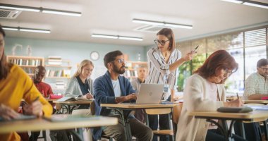 Genç öğretmen dizüstü bilgisayarla Hintli bir adamı kontrol ediyor, ona bir görevde yardım ediyor, çalışmalarına iltifat ediyor. Yetişkin Eğitim Merkezi İnsanlara Yardım Eder