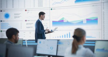 Erkek Finansal Analizci Modern İzleme Ofisi 'ndeki Büyük Dijital Ekran Raporları ve Bilgileri İnceledi. Arkasında çalışan çeşitli çalışanlar