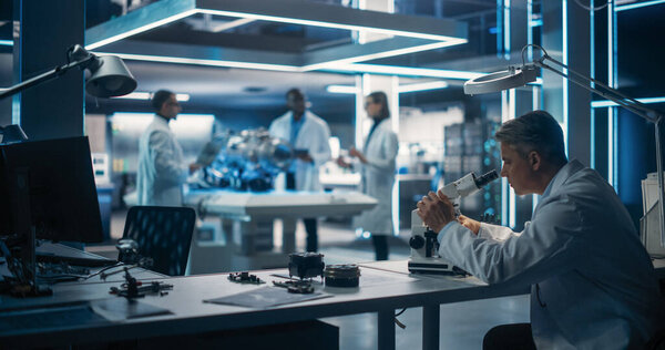 Молодая команда мультикультурных инженеров использует чертежи, планшеты, портативные компьютеры и микроскоп для анализа и обсуждения того, как работает электротурбинный двигатель