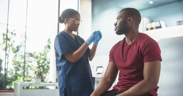 stock image African American Man Sitting In The Chair In Hospital And Getting His Chicken Pox Vaccine. Professional Black Female Nurse Is Performing Injection And