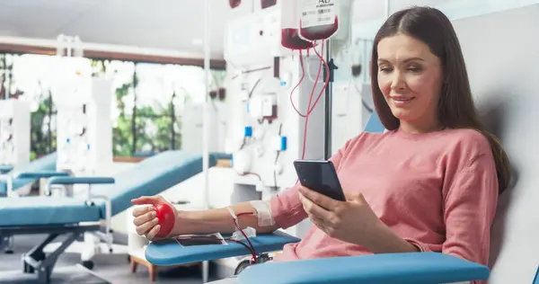 stock image Caucasian Woman Donating Blood For People In Need. Female Donor Squeezing Heart-Shaped Red Ball To Pump Blood And Chatting Online Using Smartphone