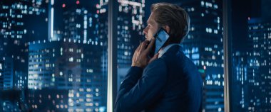 Gece geç saatlerde Gökdelenlerle Büyük Şehir Ofisi 'nde Pencerenin yanında duran şık takım elbiseli yakışıklı beyaz iş adamının arka planı. Erkek CEO konuşuyor.