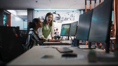 Siyah Kadın Oyun Geliştirici Masaüstü Bilgisayar kullanarak 3D modelleme yazılımı kullanıyor ve Oyun Tasarım Stüdyosu Ofisinde Asyalı Kadın Proje Yöneticisi ile konuşuyor