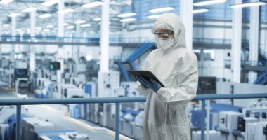 Bir üretim tesisinde tek kullanımlık koruyucu giysi, maske ve gözlük takan bir endüstri bilimcisi. Tablet Bilgisayarı Kullanılan Kadın, Potansiyel Analiz Ediliyor