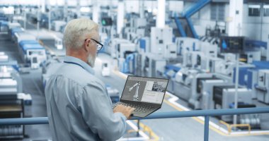 Gözlüklü yakışıklı bir orta yaşlı mühendisin portresi dizüstü bilgisayar kullanıyor ve ekipman üreten bir fabrika tesisine bakıyor.