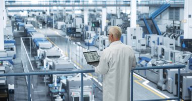 Deneyimli Endüstriyel Bilimadamı Beyaz Önlük Giyiyor, Dizüstü bilgisayar kullanıyor ve Modern Otomatik Elektronik Üretiminde Gözden Kaçan Üretim