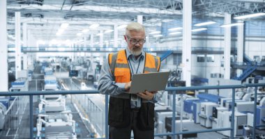 Kıdemli mühendis bir platformda duruyor, bir elektronik fabrikasında dizüstü bilgisayar kullanıyor. Makineler Bakım ve Uzman Gözlem Altından Geçiyor