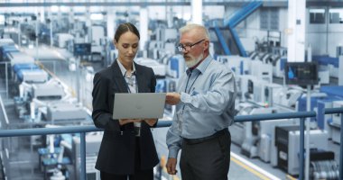 Experienced Male and Female Engineers Standing on a Platform at a Modern Industrial Factory, Using Laptop Computer and Discussing Production of Modern clipart