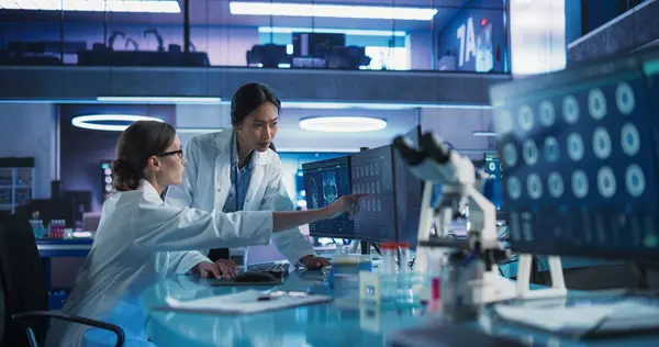 stock image Medical Research Center Laboratory: Caucasian And Asian Female Scientists Talking And Using Desktop Computer With MRI Scans Of Human Brain. Surgeon